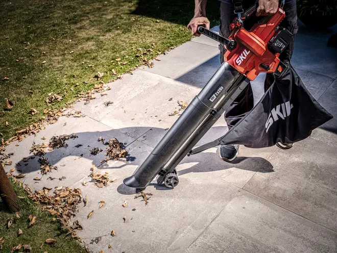 Rød SKIL rygblæser med sort opsamlingspose og langt mundstykke, vist mens den rydder blade fra en betonsti.