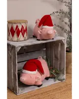 Two plush pig toys with light pink bodies, embroidered facial features, and small curly tails. Each pig is wearing a red and white Santa hat with a gold bell on the tip. The pigs are displayed on a rustic wooden crate, with one on top and one inside the crate. The setting includes a decorative drum with red diamond patterns and some pine branches for a festive touch.