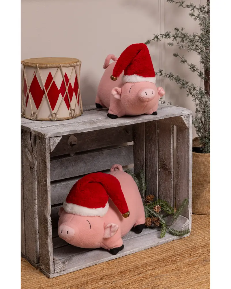 Two plush pig toys with light pink bodies, embroidered facial features, and small curly tails. Each pig is wearing a red and white Santa hat with a gold bell on the tip. The pigs are displayed on a rustic wooden crate, with one on top and one inside the crate. The setting includes a decorative drum with red diamond patterns and some pine branches for a festive touch.