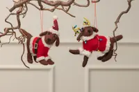 Set med två filtade hundjulprydnader, en klädd som tomte och en som ren med ljusslinga, båda i rödvit dräkt, hängande från grenar.
