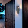 Moderne udendørs væglampe med sort cylindrisk design, monteret på hvid væg. Udsender varmt lys, perfekt til indgange eller terrasser. Vejrbestandig og stilfuld.