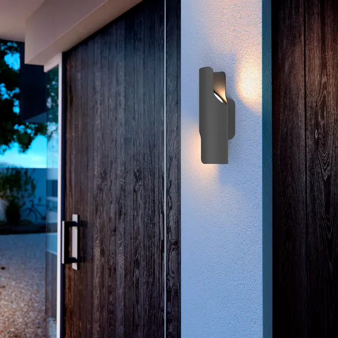 Moderne udendørs væglampe med sort cylindrisk design, monteret på hvid væg. Udsender varmt lys, perfekt til indgange eller terrasser. Vejrbestandig og stilfuld.