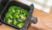 Svart airfryer-korg med flera färska broccolibuketter, placerad på en träbänk. En hand håller i handtaget, redo att luftfritera grönsakerna.