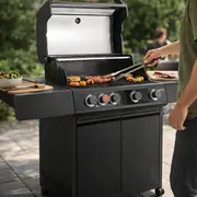 Sort gasgrill med åbent låg, hvor en person griller kød og grøntsager på terrassen.