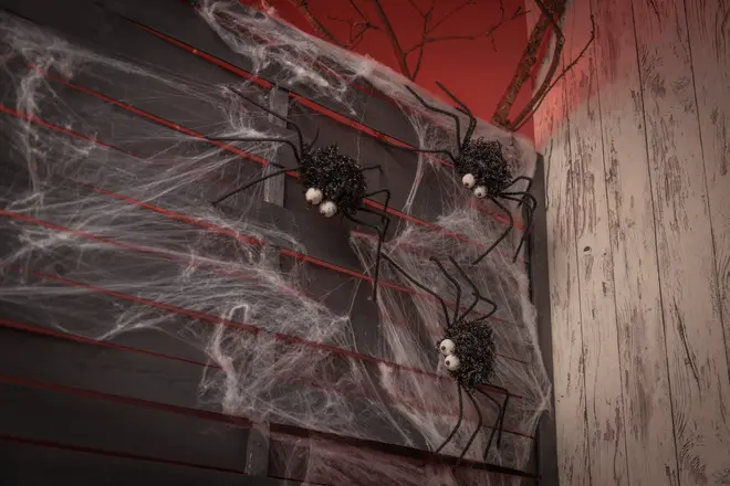 Halloween-väggdekoration med tre stora svarta spindlar med vita ögon och konstgjorda spindelnät, fästa på en mörk trävägg för en kuslig effekt.